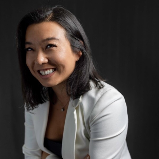 Photo of Irene Phan at three quarter angle in front of a gray background, wearing a white blazer and dark cami-top, with shoulder length bobbed hair and a big smile.