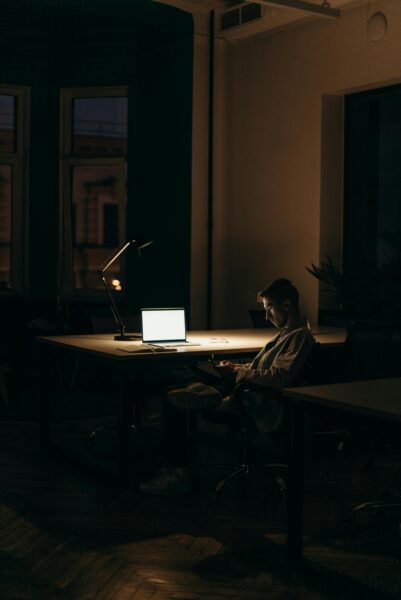 A man sits at a table in a dark room with only a pool of light on his open laptop- but he is looking at his phone.
Photo credit: cottonbro studio https://www.pexels.com/search/cottonbro%20studio/